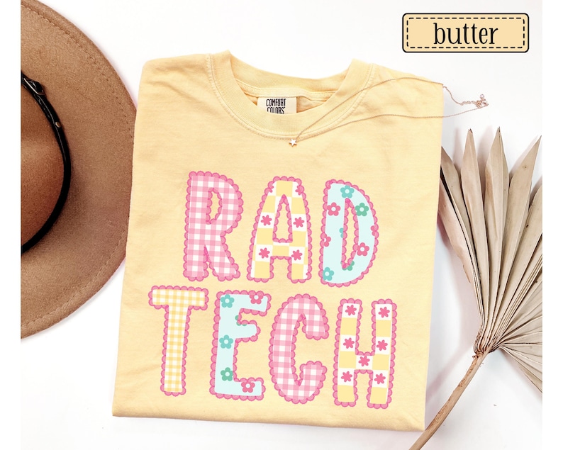 May include: A light yellow t-shirt with the words "RAD TECH" in a colorful, patchwork design. The letters are outlined in pink and blue, with floral accents. The word "butter" is in a small rectangle in the upper right corner.