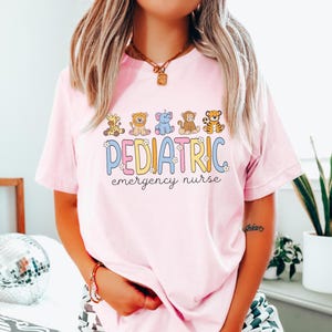 May include: Pink t-shirt with a graphic of cartoon animals and the text "PEDIATRIC emergency nurse".