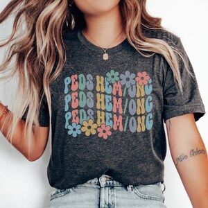 May include: A dark grey t-shirt with the text "PEDS HEM/ONG" repeated three times in a retro font with colorful flowers in between each line. The shirt is being worn by a person with long blonde hair.
