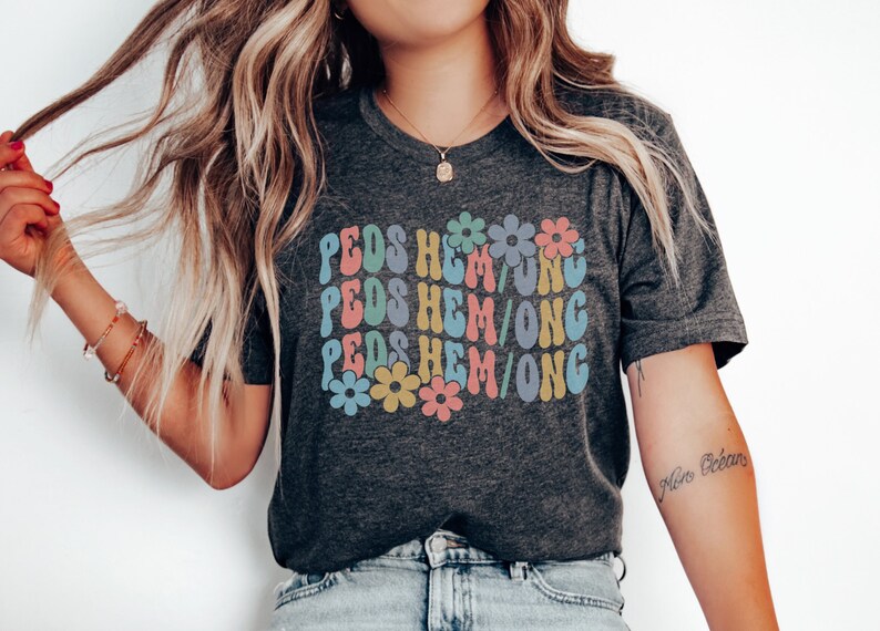 May include: A dark grey t-shirt with the text "PEDS HEM/ONG" repeated three times in a retro font with colorful flowers in between each line. The shirt is being worn by a person with long blonde hair.