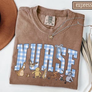 K&ouml;nnte beinhalten: Braunes T-Shirt mit dem Wort "NURSE" in blau-wei&szlig;em Vichy-Schriftzug. Das Shirt zeigt Zeichentrickfiguren unter dem Wort. Eine silberne Halskette mit einem kleinen Anh&auml;nger befindet sich auf dem Shirt.