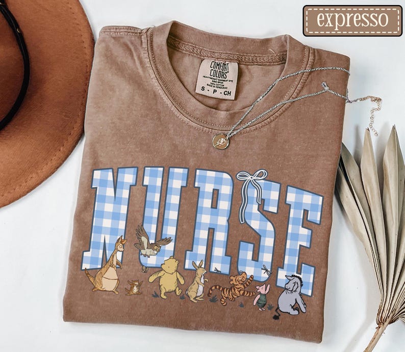 K&ouml;nnte beinhalten: Braunes T-Shirt mit dem Wort "NURSE" in blau-wei&szlig;em Vichy-Schriftzug. Das Shirt zeigt Zeichentrickfiguren unter dem Wort. Eine silberne Halskette mit einem kleinen Anh&auml;nger befindet sich auf dem Shirt.