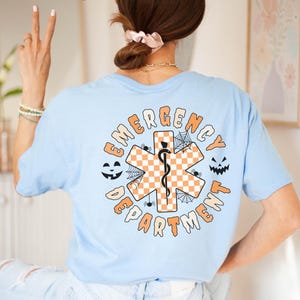 Op de afbeelding: Een lichtblauw t-shirt met een afbeelding op de achterkant met de tekst EMERGENCY DEPARTMENT. Het ontwerp bevat een oranje en wit geblokt symbool van de Ster van het Leven met een zwarte slang en staf, omringd door spinnenwebben, spinnen en pompoengezichten.