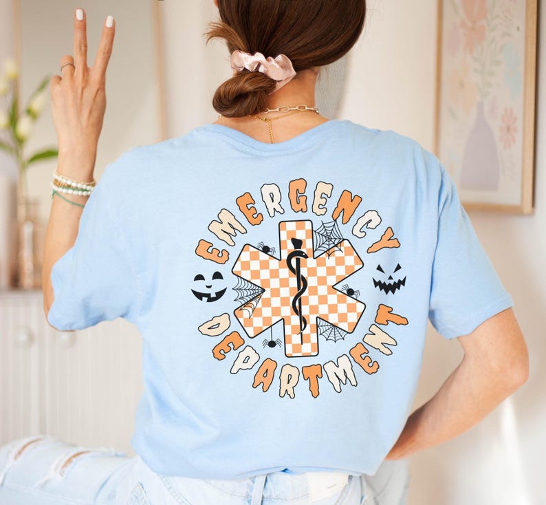 Op de afbeelding: Een lichtblauw t-shirt met een afbeelding op de achterkant met de tekst EMERGENCY DEPARTMENT. Het ontwerp bevat een oranje en wit geblokt symbool van de Ster van het Leven met een zwarte slang en staf, omringd door spinnenwebben, spinnen en pompoengezichten.