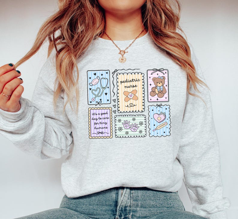 May include: Light gray sweatshirt with colorful illustrations related to pediatric nursing. The design includes a stethoscope, band-aid, teddy bear, and the text "It's a good day to care for tiny humans."