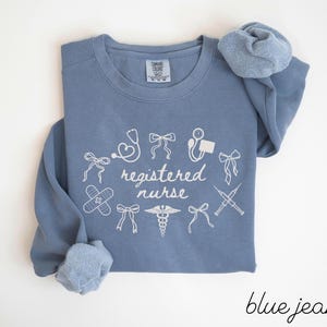 May include: A blue jean colored long-sleeve sweatshirt with the words "registered nurse" in white script. The sweatshirt has white medical-themed graphics, including a stethoscope, band-aid, and syringe.