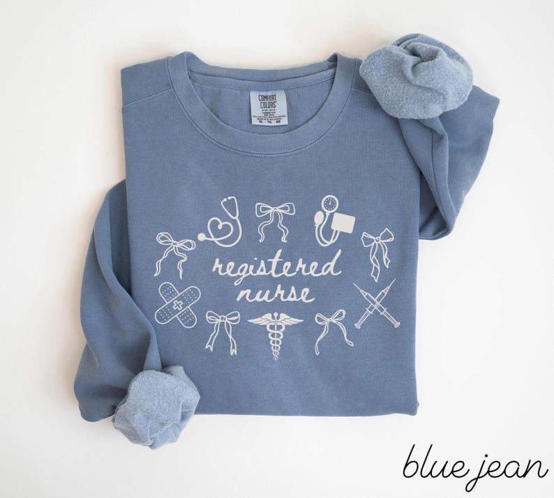 May include: A blue jean colored long-sleeve sweatshirt with the words "registered nurse" in white script. The sweatshirt has white medical-themed graphics, including a stethoscope, band-aid, and syringe.