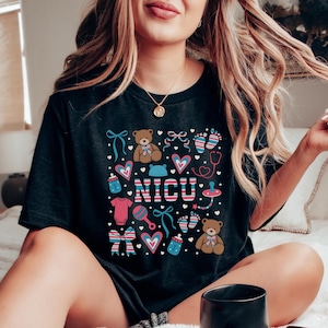 May include: Black t-shirt with a colorful design featuring a teddy bear, a baby bottle, a rattle, and the word "NICU".