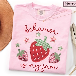 K&ouml;nnte beinhalten: Ein hellrosa T-Shirt mit einer zentralen Grafik aus drei Erdbeeren mit rot-gr&uuml;nem Vichy-Muster. Der Text "behavior is my jam" ist in einer roten Schreibschrift gedruckt. Kleine Vichy-Sterne umgeben die Erdbeeren.