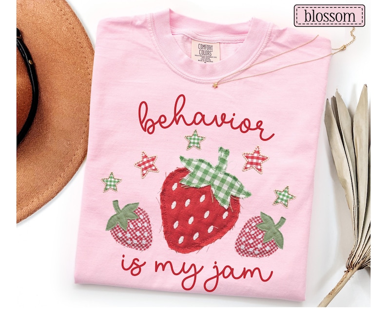 K&ouml;nnte beinhalten: Ein hellrosa T-Shirt mit einer zentralen Grafik aus drei Erdbeeren mit rot-gr&uuml;nem Vichy-Muster. Der Text "behavior is my jam" ist in einer roten Schreibschrift gedruckt. Kleine Vichy-Sterne umgeben die Erdbeeren.