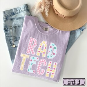May include: A lilac-colored t-shirt with the words "RAD TECH" in a patchwork design. The letters feature gingham and floral patterns. A pair of blue jeans and a tan hat are also in the image. The shirt is labeled "orchid".