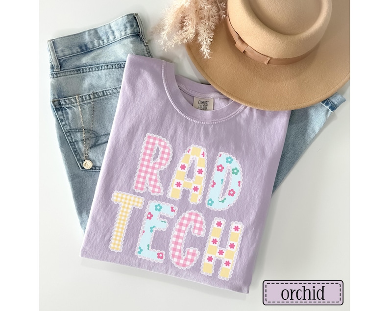 May include: A lilac-colored t-shirt with the words "RAD TECH" in a patchwork design. The letters feature gingham and floral patterns. A pair of blue jeans and a tan hat are also in the image. The shirt is labeled "orchid".