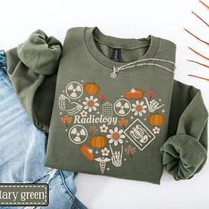 May include: A military green crewneck sweatshirt featuring a heart-shaped graphic composed of pumpkins, flowers, radiation symbols, skeleton hands, a clipboard, a slice of pie, and an X-ray, with the word Radiology printed in the center.