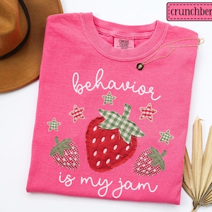 K&ouml;nnte beinhalten: Ein leuchtend pinkfarbenes T-Shirt mit einer zentralen Grafik aus drei Erdbeeren mit gr&uuml;n-roten Vichy-Stoffmustern. Der Text "behavior is my jam" ist in wei&szlig;er Schreibschrift gedruckt. F&uuml;nf kleine Vichy-Sterne umgeben die Erdbeeren.