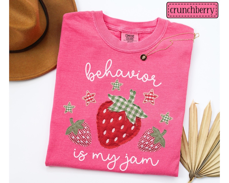K&ouml;nnte beinhalten: Ein leuchtend pinkfarbenes T-Shirt mit einer zentralen Grafik aus drei Erdbeeren mit gr&uuml;n-roten Vichy-Stoffmustern. Der Text "behavior is my jam" ist in wei&szlig;er Schreibschrift gedruckt. F&uuml;nf kleine Vichy-Sterne umgeben die Erdbeeren.
