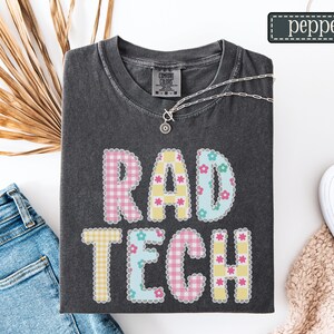 May include: Dark gray t-shirt with the words "RAD TECH" in a patchwork design. The letters are in pink, yellow, and blue with floral and gingham patterns. A silver necklace with a pendant is draped on the shirt.
