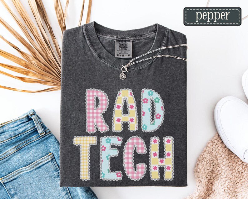 May include: Dark gray t-shirt with the words "RAD TECH" in a patchwork design. The letters are in pink, yellow, and blue with floral and gingham patterns. A silver necklace with a pendant is draped on the shirt.