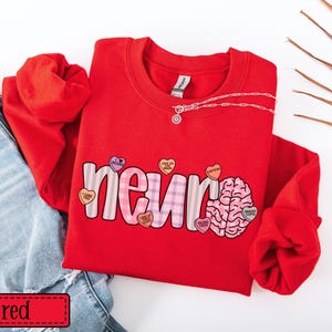May include: A red sweatshirt with the word "neuron" in a playful font, featuring a brain graphic and heart-shaped candies. A silver chain necklace is draped across the top. The word "red" is in a red rectangle at the bottom.