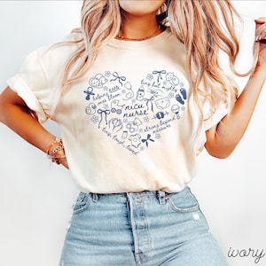 May include: Ivory-colored t-shirt featuring a navy blue heart-shaped design with baby-related illustrations and text. The text includes "nicu nurse," "tiny but mighty," and "helping little ones bloom." The word "ivory" is printed at the bottom.