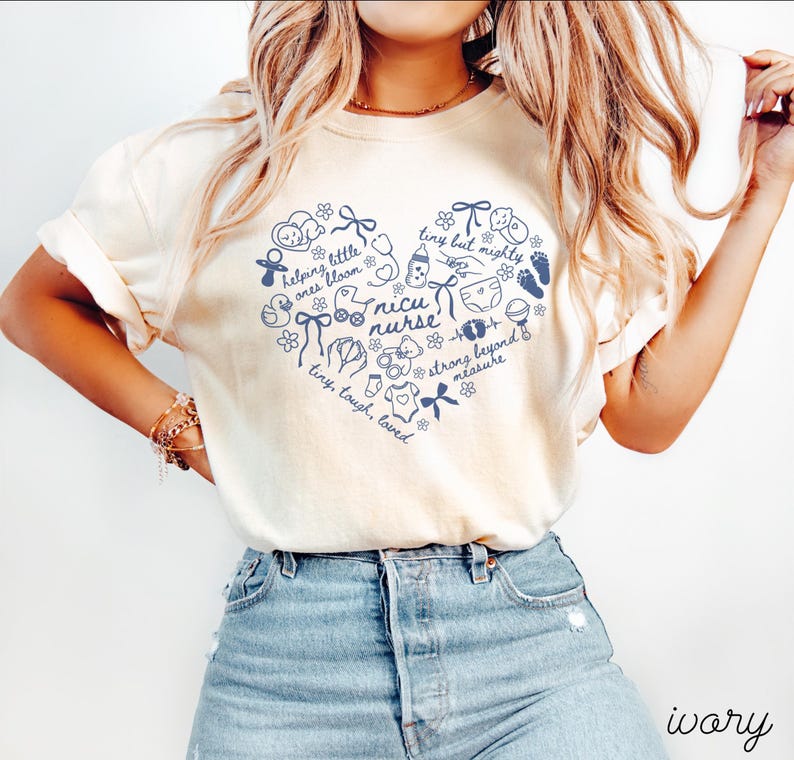 May include: Ivory-colored t-shirt featuring a navy blue heart-shaped design with baby-related illustrations and text. The text includes "nicu nurse," "tiny but mighty," and "helping little ones bloom." The word "ivory" is printed at the bottom.