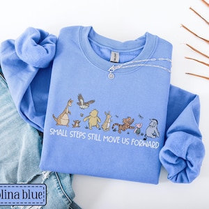 May include: A Carolina blue sweatshirt with a graphic of Winnie the Pooh characters and the text "Small steps still move us forward." A silver necklace is on the shirt. The sweatshirt is paired with blue jeans.