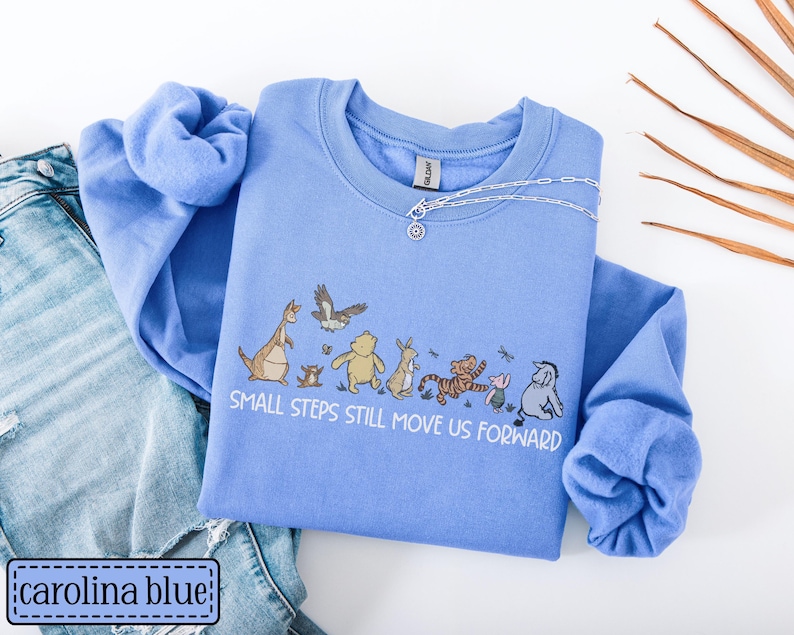 May include: A Carolina blue sweatshirt with a graphic of Winnie the Pooh characters and the text "Small steps still move us forward." A silver necklace is on the shirt. The sweatshirt is paired with blue jeans.