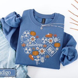 May include: A blue crewneck sweatshirt featuring a heart-shaped graphic composed of pumpkins, daisies, autumn leaves, radiation symbols, a clipboard, a slice of pie, and skeletal hand illustrations with the word Radiology printed in the center.