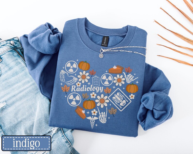 May include: A blue crewneck sweatshirt featuring a heart-shaped graphic composed of pumpkins, daisies, autumn leaves, radiation symbols, a clipboard, a slice of pie, and skeletal hand illustrations with the word Radiology printed in the center.