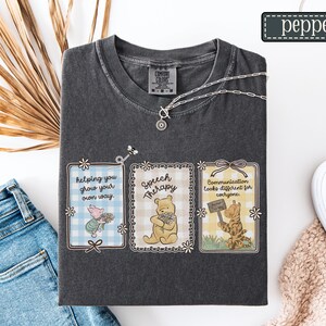 May include: Dark gray t-shirt with three framed illustrations featuring cartoon characters and text. The frames read "Helping you grow your own way," "Speech Therapy," and "Communication looks different for everyone." A silver necklace is draped across the shirt.