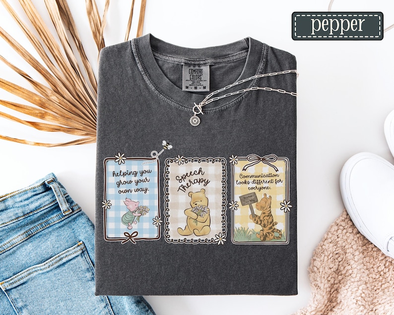 May include: Dark gray t-shirt with three framed illustrations featuring cartoon characters and text. The frames read "Helping you grow your own way," "Speech Therapy," and "Communication looks different for everyone." A silver necklace is draped across the shirt.