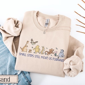 May include: A sand-coloured sweatshirt with a crew neck and the phrase "SMALL STEPS STILL MOVE US FORWARD" printed in blue. The design features cartoon characters. A silver necklace with a pendant is visible.