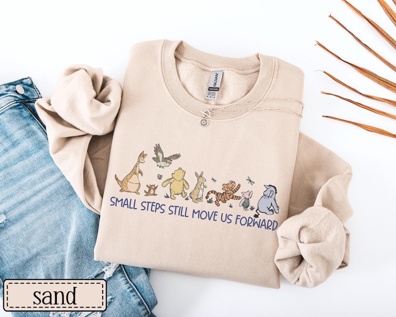 May include: A sand-coloured sweatshirt with a crew neck and the phrase "SMALL STEPS STILL MOVE US FORWARD" printed in blue. The design features cartoon characters. A silver necklace with a pendant is visible.