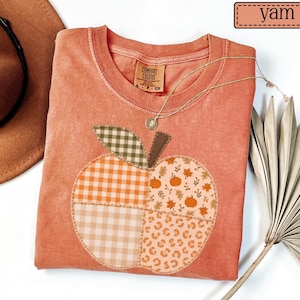 May include: A yam-coloured crew neck t-shirt featuring a large apple-shaped appliqué on the front. The apple is divided into four sections with different patterns: orange gingham, light beige gingham, autumn leaves with pumpkins, and leopard print.