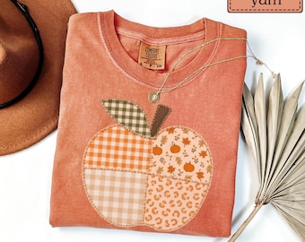 May include: A yam-colored crew neck t-shirt featuring a large apple-shaped applique on the front. The apple is divided into four sections with different patterns: orange gingham, light beige gingham, autumn leaves with pumpkins, and leopard print.