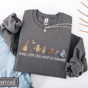 May include: Charcoal grey sweatshirt featuring the phrase "SMALL STEPS STILL MOVE US FORWARD" and a cartoon illustration of Winnie the Pooh characters. A silver necklace is draped across the sweatshirt. The sweatshirt is folded next to a pair of blue jeans.