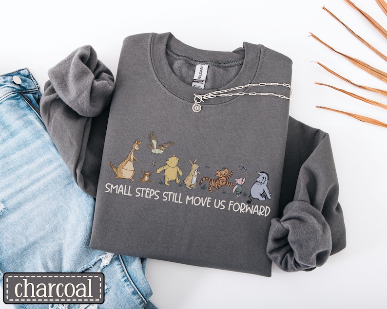 May include: Charcoal grey sweatshirt featuring the phrase "SMALL STEPS STILL MOVE US FORWARD" and a cartoon illustration of Winnie the Pooh characters. A silver necklace is draped across the sweatshirt. The sweatshirt is folded next to a pair of blue jeans.