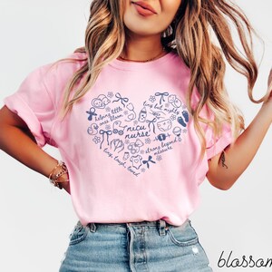 May include: A light pink t-shirt with a navy blue heart-shaped design. The heart contains the words "nicu nurse" and other text and illustrations related to babies. The word "blossom" is written in cursive at the bottom.