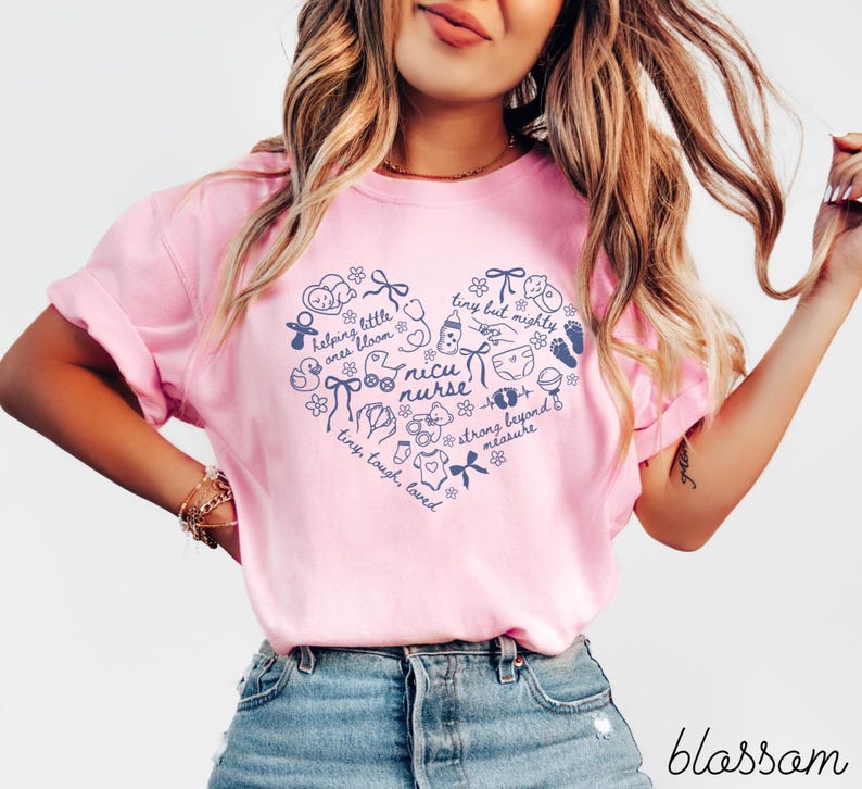 May include: A light pink t-shirt with a navy blue heart-shaped design. The heart contains the words "nicu nurse" and other text and illustrations related to babies. The word "blossom" is written in cursive at the bottom.