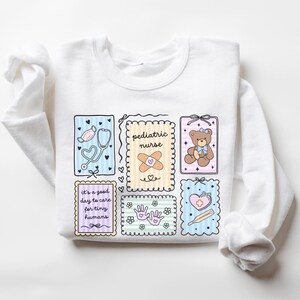 May include: White long-sleeved sweatshirt featuring colorful illustrations related to pediatric care. The design includes a stethoscope, teddy bear, bandage, and the text "pediatric nurse" and "it's a good day to care for tiny humans."