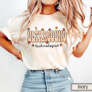 Puede incluir: Camiseta color marfil con la palabra "ULTRASOUND" en un dise&ntilde;o a cuadros y "technologist" debajo. El dise&ntilde;o incluye hojas de oto&ntilde;o y bellotas. La camiseta es de estilo casual.