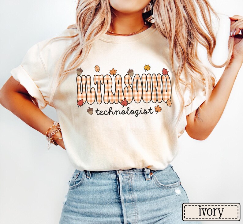 Puede incluir: Camiseta color marfil con la palabra "ULTRASOUND" en un dise&ntilde;o a cuadros y "technologist" debajo. El dise&ntilde;o incluye hojas de oto&ntilde;o y bellotas. La camiseta es de estilo casual.