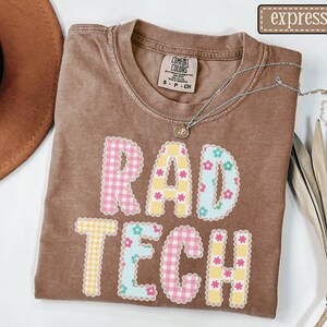 May include: A brown t-shirt with the words "RAD TECH" in a colorful, patterned design. The letters are made of pink and yellow gingham, and blue floral patterns. A silver necklace with a small pendant rests on the shirt.