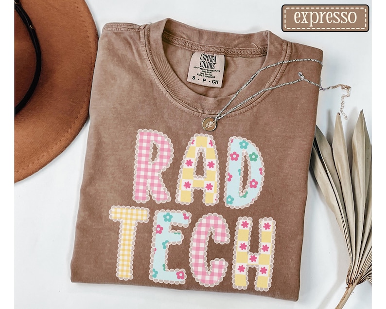 May include: A brown t-shirt with the words "RAD TECH" in a colorful, patterned design. The letters are made of pink and yellow gingham, and blue floral patterns. A silver necklace with a small pendant rests on the shirt.