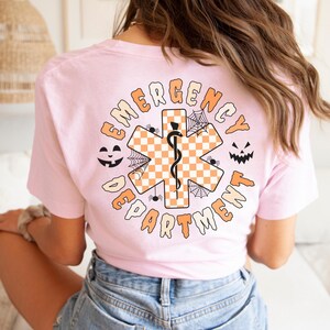 Op de afbeelding: Een lichtroze t-shirt met een achteropdruk van een oranje en wit geblokt Ster van Leven-symbool met een zwarte slang en staf. Het ontwerp bevat zwarte spinnenwebben, twee pompoengezichten en de tekst EMERGENCY DEPARTMENT.