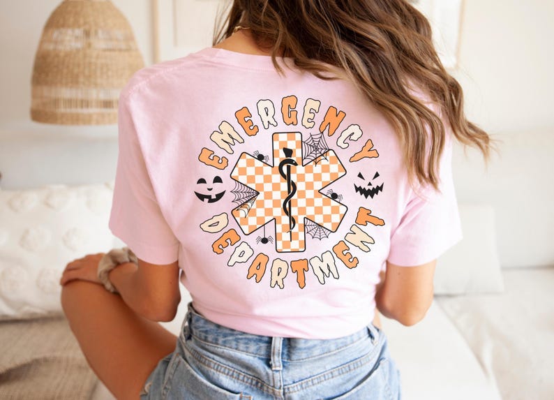Op de afbeelding: Een lichtroze t-shirt met een achteropdruk van een oranje en wit geblokt Ster van Leven-symbool met een zwarte slang en staf. Het ontwerp bevat zwarte spinnenwebben, twee pompoengezichten en de tekst EMERGENCY DEPARTMENT.