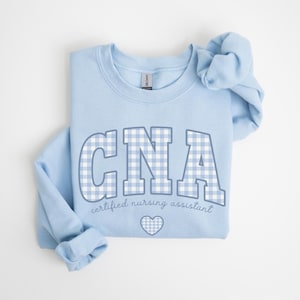 May include: Light blue sweatshirt with a white and blue gingham pattern spelling "CNA" with the text "certified nursing assistant" below. A small white heart is centered below the text.