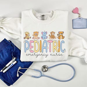 May include: White sweatshirt with a graphic of cartoon animals and the text "PEDIATRIC emergency nurse". The sweatshirt is laid out on a white surface with a stethoscope, scrubs, pens, a badge holder, and a smartwatch.