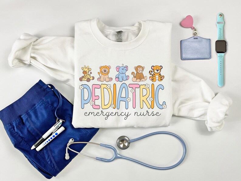 May include: White sweatshirt with a graphic of cartoon animals and the text "PEDIATRIC emergency nurse". The sweatshirt is laid out on a white surface with a stethoscope, scrubs, pens, a badge holder, and a smartwatch.