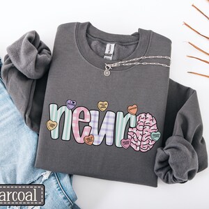 May include: Charcoal gray sweatshirt with the word "neur" in pastel colors and a brain graphic. The word "neur" is decorated with heart-shaped candies. A silver necklace is draped across the sweatshirt. The word "charcoal" is in a black rectangle.