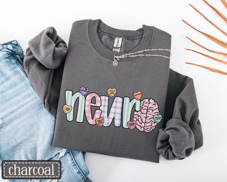 May include: Charcoal gray sweatshirt with the word "neur" in pastel colors and a brain graphic. The word "neur" is decorated with heart-shaped candies. A silver necklace is draped across the sweatshirt. The word "charcoal" is in a black rectangle.